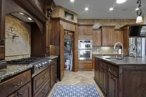 Kitchen featuring stainless steel refrigerator, TV, gas cooktop, double oven, light tile floors, granite-like countertops, dark brown cabinetry, and kitchen island sink