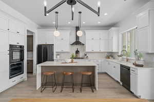 Kitchen with a breakfast bar, a kitchen island, natural light, refrigerator, exhaust hood, stainless steel dishwasher, double oven, light hardwood flooring, light countertops, pendant lighting, and white cabinetry