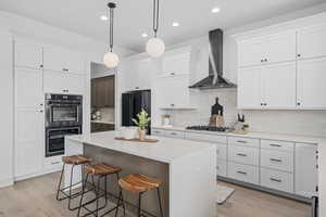 Kitchen with a center island, a kitchen breakfast bar, refrigerator, gas stovetop, ventilation hood, stainless steel double oven, light hardwood floors, light countertops, pendant lighting, and white cabinetry