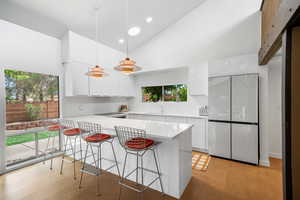 Kitchen featuring a kitchen bar, vaulted ceiling, natural light, refrigerator, pendant lighting, light floors, light countertops, and white cabinetry