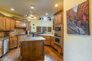 Kitchen with backsplash, an island with sink, light countertops, brown cabinets, custom exhaust hood, and appliances with stainless steel finishes