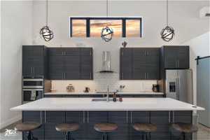 Kitchen featuring pendant lighting, dark brown cabinets, appliances with stainless steel finishes, wall chimney range hood, light countertops, backsplash, and a center island