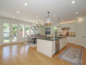 Kitchen featuring pendant lighting, an island with sink, light hardwood flooring, dark countertops, french doors, stainless steel dishwasher, backsplash, ornamental molding, and white cabinetry