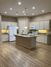 Kitchen featuring granite-like countertops, kitchen island with sink, white appliances, hanging light fixtures,, and light LVP floors