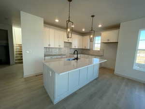 Kitchen with hanging light fixtures, kitchen island with sink, backsplash, light countertops, white cabinetry, and hardwood flooring