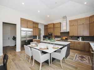 Kitchen featuring wall chimney exhaust hood, built in appliances, brown cabinets, light countertops, light hardwood flooring, and backsplash