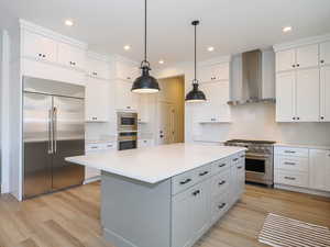 Kitchen featuring wall chimney exhaust hood, built in appliances, a kitchen island, and light wood-type flooring