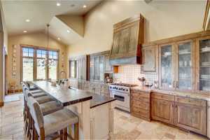Kitchen with a kitchen island, lofted ceiling, light stone countertops, brown cabinets, high end stainless steel range, premium range hood, backsplash, a high ceiling, and light tile floors