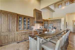 Kitchen with backsplash, high end stainless steel range, brown cabinets, light tile floors, premium range hood, and a high ceiling
