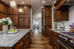 Kitchen with stainless steel appliances, pendant lighting, dark brown cabinetry, dark hardwood floors, custom exhaust hood, light stone countertops, and backsplash