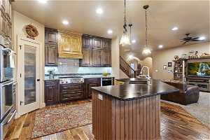 Kitchen with appliances with stainless steel finishes, custom range hood, pendant lighting, dark brown cabinets, light hardwood flooring, backsplash, and ceiling fan