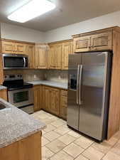 Kitchen with a textured ceiling, backsplash, brown cabinets, light countertops, stainless steel appliances, and light tile flooring