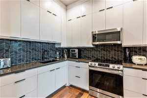 Kitchen featuring backsplash, wood-type flooring, white cabinetry, stainless steel appliances, and dark countertops