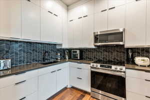 Kitchen featuring backsplash, wood-type flooring, white cabinetry, stainless steel appliances, and dark countertops