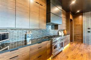 Kitchen featuring wall chimney range hood, double oven range, dark countertops, brown cabinets, light hardwood flooring, and backsplash