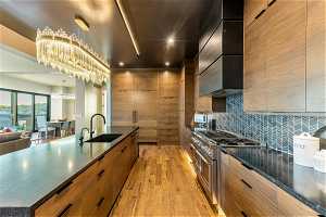 Kitchen featuring double oven range, backsplash, brown cabinets, light hardwood flooring, wall chimney exhaust hood, and dark countertops