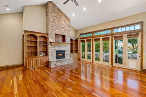 Unfurnished living room featuring a stone fireplace, vaulted ceiling high, ceiling fan, built in shelves, and hardwood floors