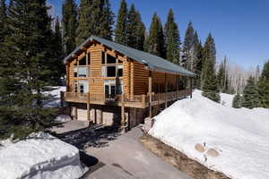 View of snowy exterior with log exterior, stone siding, driveway, a garage, and a view of trees