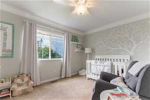 Bedroom with ceiling fan, ornamental molding, a crib, and light carpet