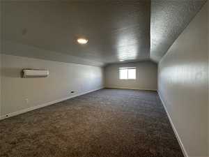 Additional living space with a textured ceiling, vaulted ceiling, an AC wall unit, and dark colored carpet