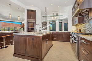 Kitchen with light stone countertops, wall chimney range hood, pendant lighting, an island with sink, and double oven range