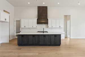 Kitchen with custom exhaust hood, light wood-style floors, a center island with sink, light countertops, and white cabinetry