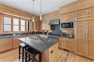 Kitchen featuring built in appliances, wall chimney exhaust hood, a kitchen island, light hardwood / wood-style floors, and decorative light fixtures