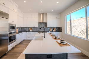 Kitchen with white cabinetry, wall chimney exhaust hood, built in appliances, a center island with sink, and dark hardwood / wood-style floors