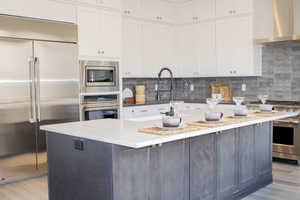 Kitchen featuring a center island, wall chimney range hood, white cabinets, built in appliances, and backsplash