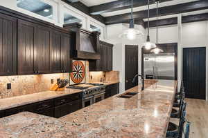 Kitchen with beam ceiling, a breakfast bar, hanging light fixtures, high end appliances, and wall chimney exhaust hood