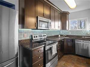 Kitchen with dark stone counters, dark brown cabinets, stainless steel appliances, backsplash, and dark tile flooring