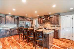 Kitchen with stainless steel appliances, DOUBLE OVEN, premium range hood, a kitchen bar, light hardwood style floors, and a kitchen island with sink