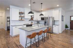 Kitchen with appliances with stainless steel finishes, white cabinets, a center island with sink, decorative light fixtures, and light wood-type flooring