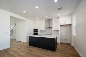 Kitchen featuring wall chimney range hood, a kitchen island with sink, stainless steel double oven, dark hardwood / wood-style floors, and white cabinetry