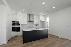 Kitchen featuring wall chimney exhaust hood, a center island with sink, sink, double oven, and white cabinetry