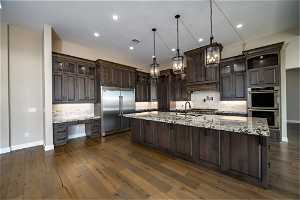 Kitchen with appliances with stainless steel finishes, backsplash, dark wood-type flooring, decorative light fixtures, and a kitchen island with sink