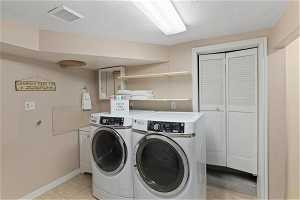 The clothes washing area features a washing machine and clothes dryer, cabinets, a textured ceiling, and light tile flooring—access to the utility room.