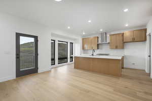 Kitchen with light hardwood / wood-style flooring, wall chimney exhaust hood, a kitchen island with sink, and sink