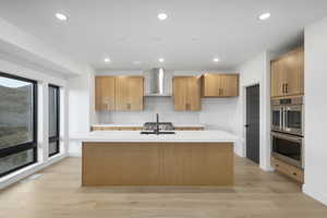 Kitchen featuring an island with sink, double oven, light hardwood / wood-style floors, wall chimney exhaust hood, and decorative backsplash