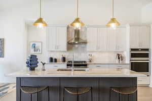 Kitchen featuring double oven, wall chimney range hood, light stone counters, white cabinetry, and light hardwood / wood-style flooring. Note: This Is Not The Subject Property Itself