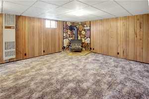 Basement with a drop ceiling, a wood stove, wood walls, and carpet floors