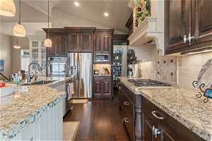 Kitchen featuring dark brown cabinets, hanging light fixtures, vaulted ceiling, stainless steel appliances, and tasteful backsplash