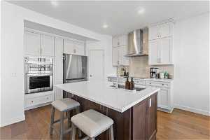 Kitchen featuring wall chimney exhaust hood, backsplash, stainless steel appliances, a center island with sink, and hardwood / wood-style flooring