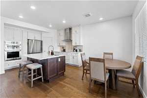 Kitchen featuring sink, wall chimney range hood, hardwood / wood-style floors, stainless steel appliances, and a kitchen island with sink