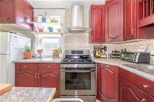 Kitchen featuring gas stove, wall chimney range hood, white fridge, and backsplash