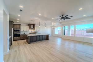 Kitchen featuring decorative light fixtures, light wood-type flooring, appliances with stainless steel finishes, a kitchen island with sink, and wall chimney range hood