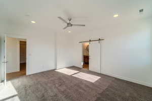 Unfurnished bedroom featuring carpet flooring, ensuite bathroom, a barn door, and ceiling fan