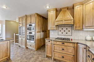 Kitchen with stainless steel double oven, backsplash, premium range hood, and light tile floors