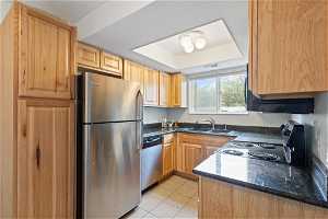 Kitchen featuring a tray ceiling, stainless steel appliances, light tile floors, sink, and dark stone countertops