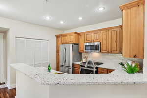 Kitchen featuring dark wood-type flooring, light stone counters, kitchen peninsula, backsplash, and appliances with stainless steel finishes